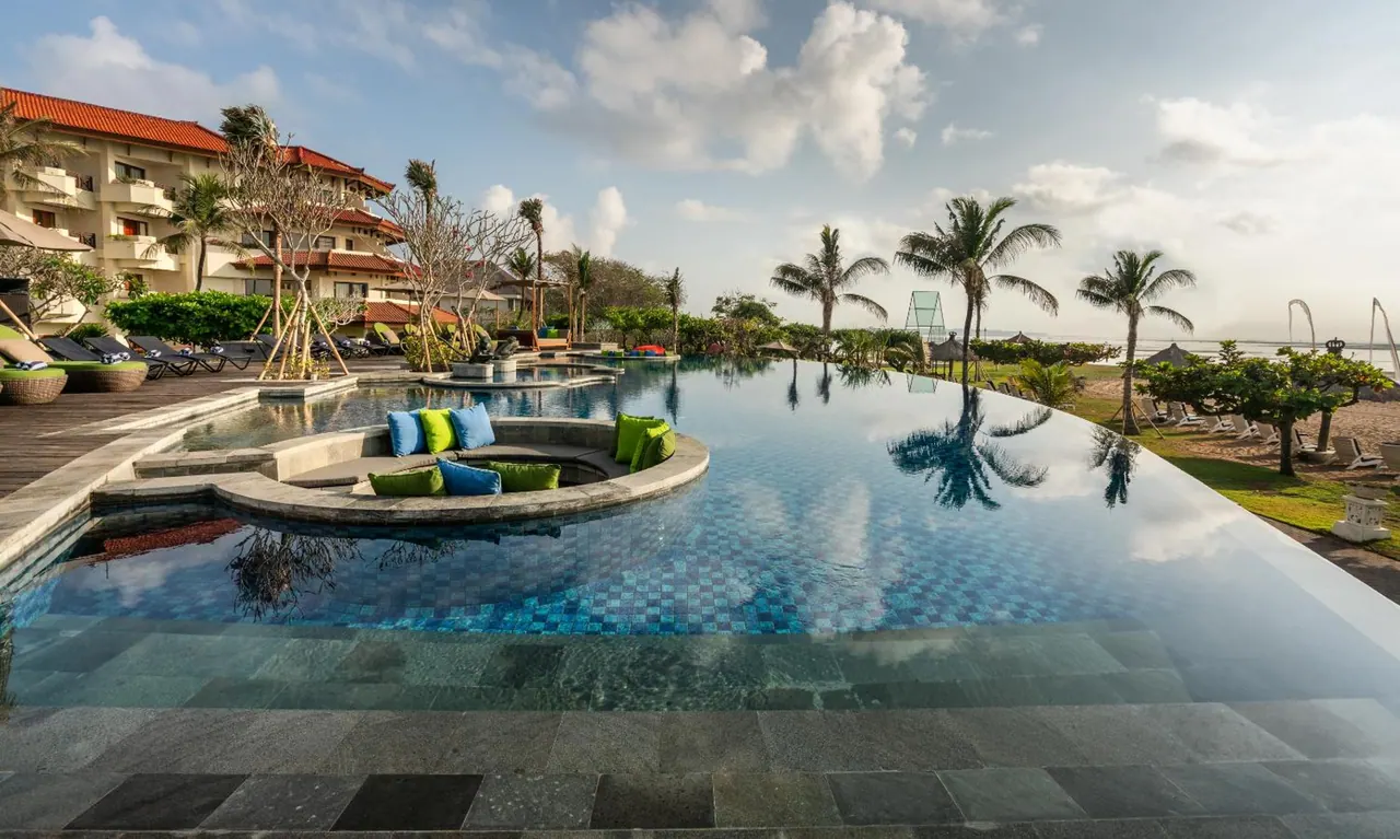An infinity-edge swimming pool with built-in cushioned seating, palm trees, sun loungers and a sandy beach with the sea beyond.