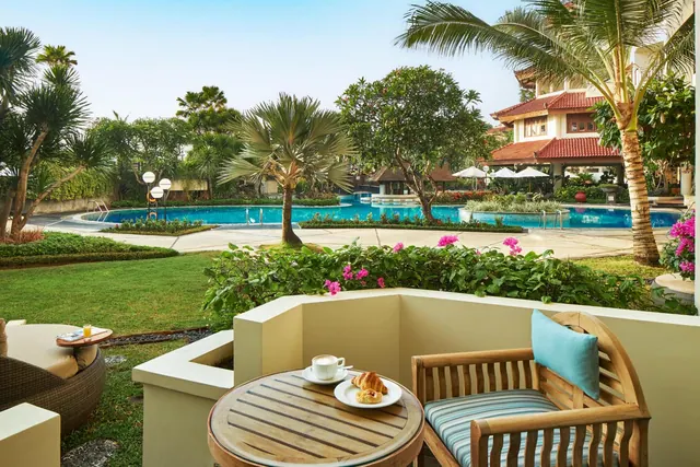 A private balcony with a wooden table and chair overlooks a large swimming pool, lawn and tropical gardens, with a coffee cup and pastries on the table.