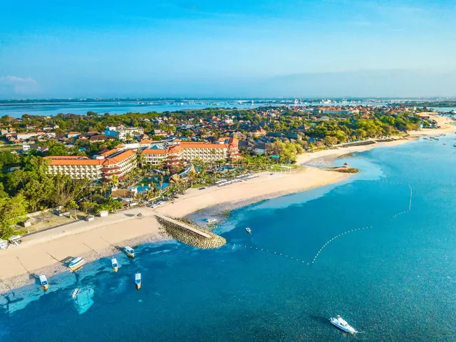Aerial view of a beachfront hotel complex with a sandy shoreline, a stone breakwater, moored boats and clear blue water.