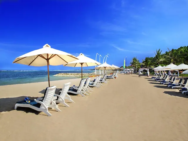 A sandy beach with rows of white sun loungers and cream parasols facing the sea under a clear blue sky.