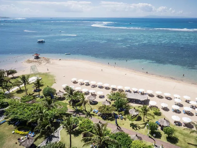Aerial view of a sandy beach with rows of white parasols and sun loungers, the sea and a reef beyond, and palm trees along the edge.