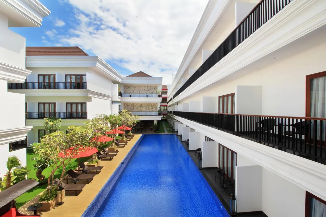 A long swimming pool runs through a courtyard lined with sun loungers, red umbrellas and white hotel buildings with balconies.