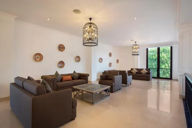 A large lounge with brown sofas and armchairs, a birdcage chandelier, and a glass door opening onto a balcony with trees outside.