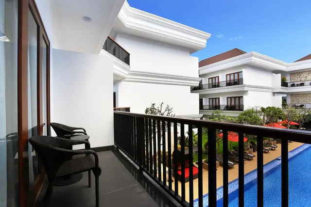 A private balcony with two wicker chairs and a small round table overlooks a swimming pool with sun loungers, red umbrellas and garden greenery.