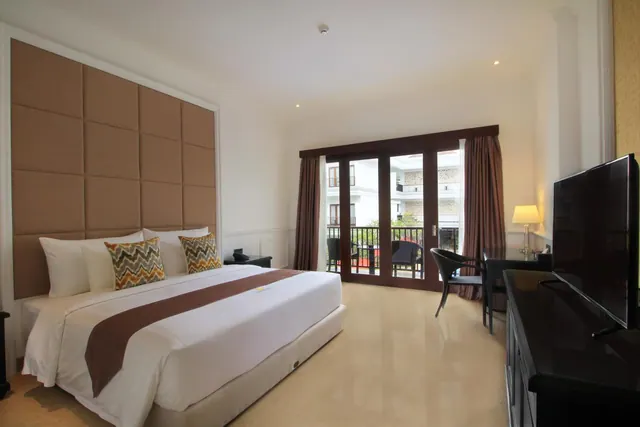A bedroom with a king bed under a tall padded headboard, a desk and chair, a flat-screen television, and glass doors opening onto a balcony with chairs and a view of a neighbouring building.
