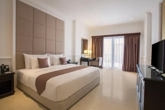 A bedroom with a king bed against an upholstered headboard wall, a desk with a chair, a wall-mounted television and sheer-curtained glass doors.