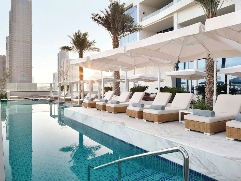 A rooftop pool with white sun loungers and umbrellas, with tall city towers visible in the background.