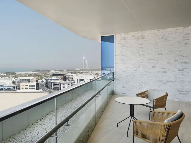 A private balcony with two woven chairs and a round table overlooks city buildings and the Burj Al Arab in the distance.