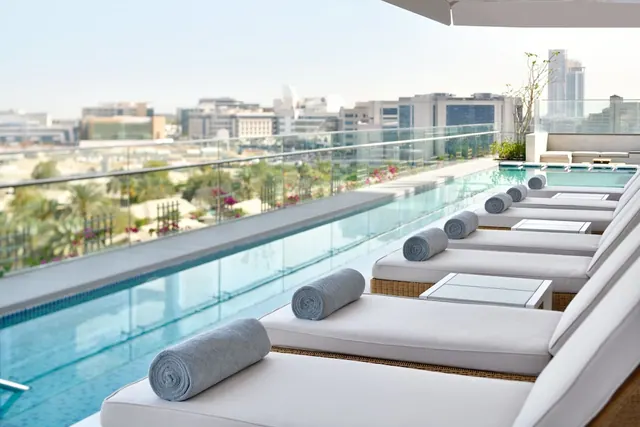 A rooftop pool with white sun loungers and rolled grey towels, overlooking a city skyline and green trees below.