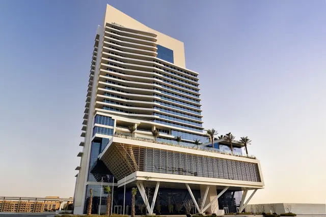 A low-angle view of a tall white hotel tower with a louvred lower block, palm trees on a rooftop terrace and a clear sky behind it.