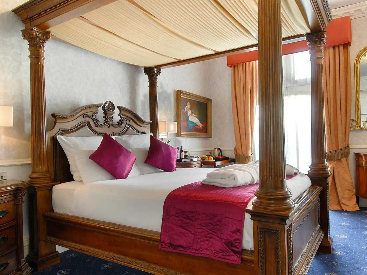A four-poster bed with a carved wooden headboard, magenta cushions and a magenta throw stands in a bedroom with a framed painting, a bedside tray and orange curtains at the window.