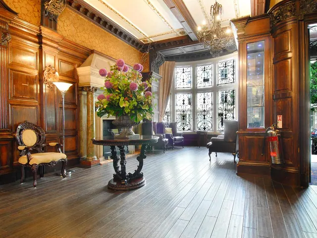 A lobby with dark wood panelling, a chandelier, a floral arrangement on a round table, armchairs, and tall windows with ornate iron grilles.