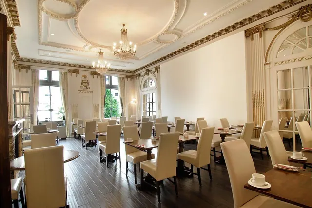 A dining room with cream leather chairs and dark wood tables, lit by chandeliers beneath an ornate moulded ceiling.