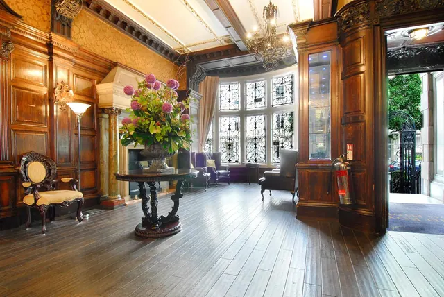 A lobby with dark wood panelling, a chandelier, a flower arrangement on a round table, armchairs and a fire extinguisher beside an open doorway to the street.
