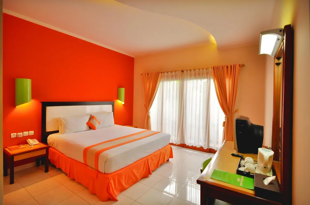 A bedroom with an orange accent wall, a double bed with an orange bed skirt, a nightstand with a telephone, and a desk with a television and kettle.