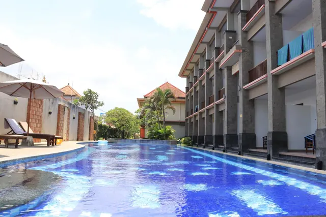 A long blue swimming pool runs past sun loungers under umbrellas, with a multi-storey hotel building with balconies on the right.