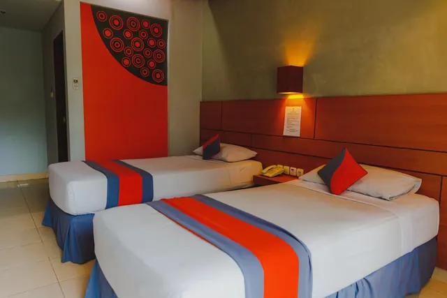 Two twin beds with red and blue runners sit against a wooden headboard beside a red accent wall with a circular pattern.