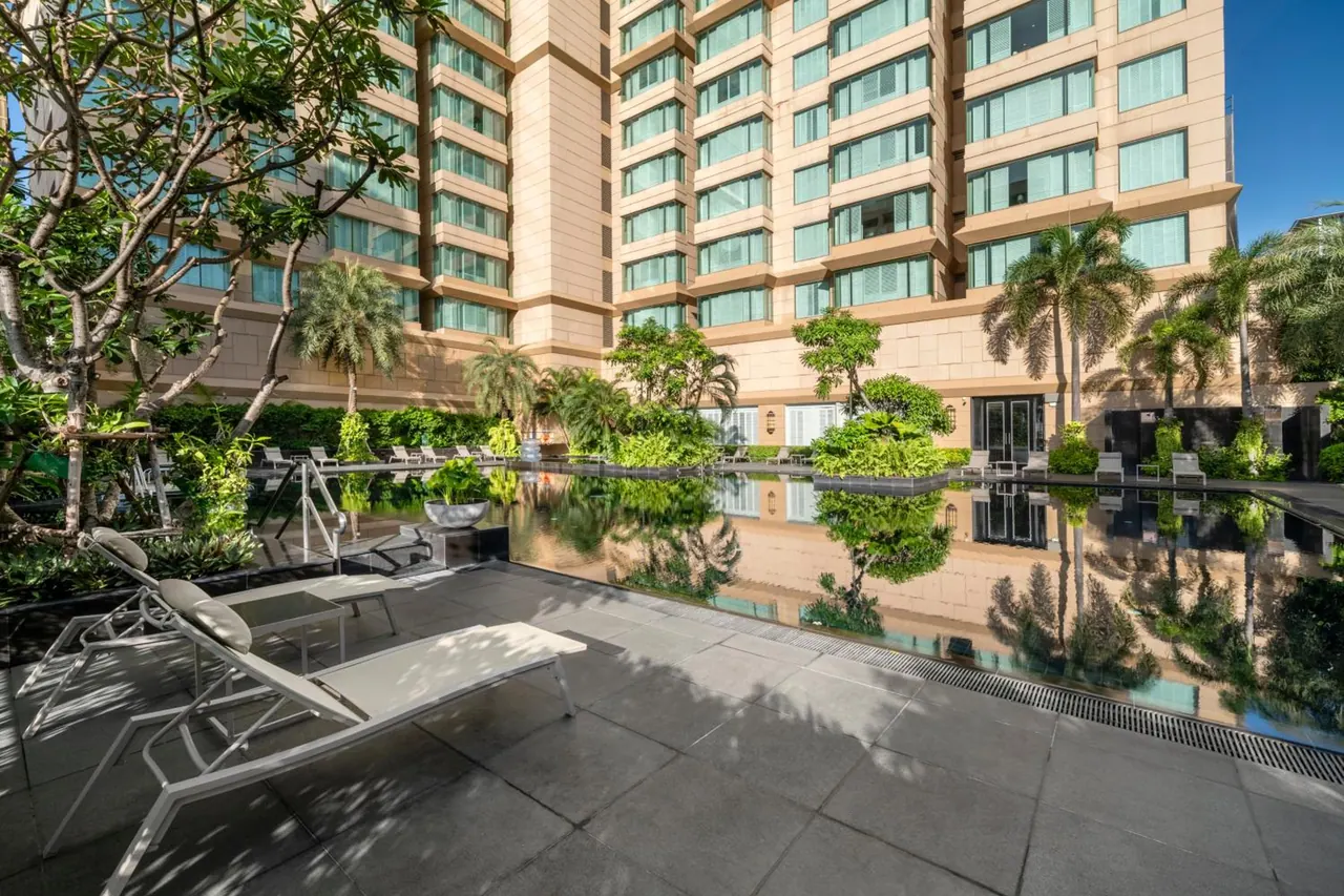 A pool with sun loungers beside a tall hotel building, with trees and palms reflected in the still water.