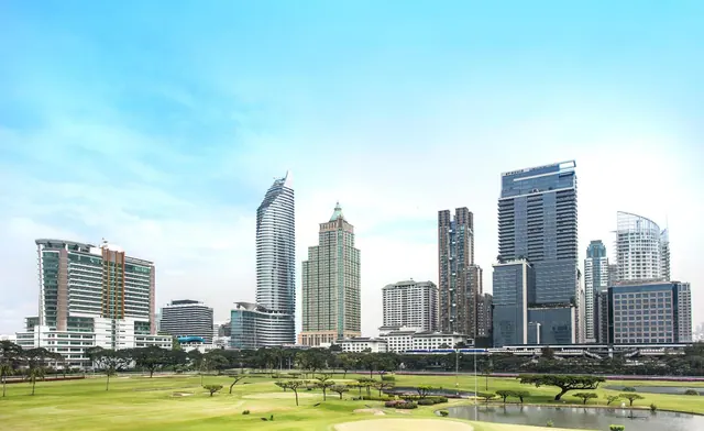 A view across a green golf course and a small pond toward a cluster of high-rise towers under a blue sky.
