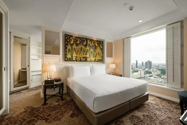 A bedroom with a made king bed, a gold-painted Thai-style wall artwork, and a large window overlooking the city skyline.