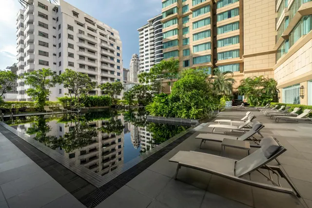 A long rectangular swimming pool reflects surrounding high-rise buildings and greenery, with grey sun loungers lined along the tiled deck.