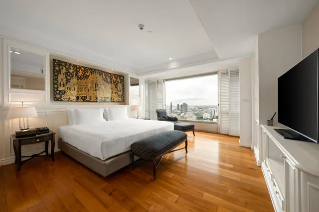 A bedroom with a king bed, a bench at the foot of the bed, an armchair, a wall tapestry, a wall-mounted television and a window looking out over city towers.