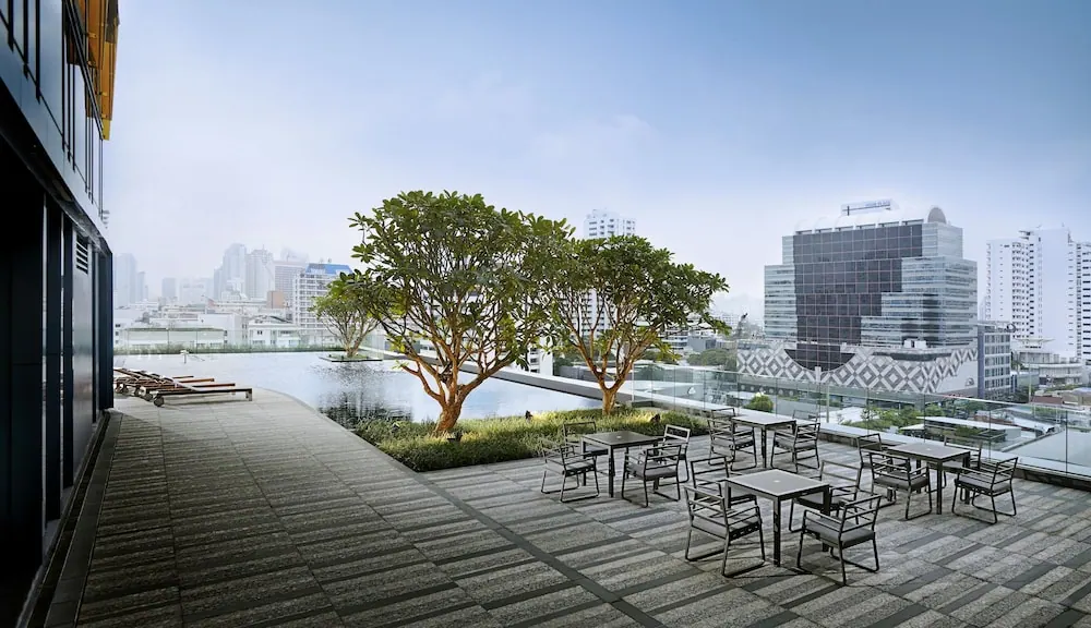 A rooftop terrace with outdoor tables and chairs, sun loungers at left, a body of water, trees and city towers beyond.