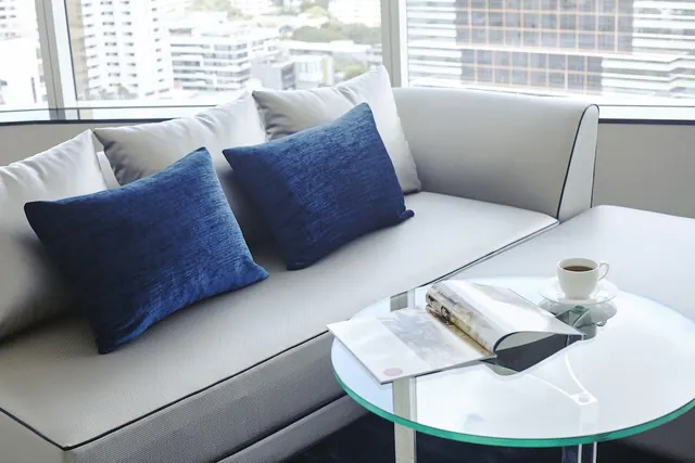 A grey sofa with blue velvet cushions sits beside a round glass side table holding a coffee cup and magazines, with high-rise buildings visible through the window.