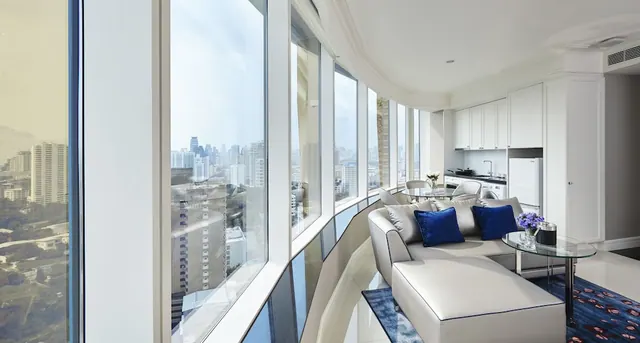 A living area with a light grey sofa, blue cushions and a small glass side table beside curved floor-to-ceiling windows looking out over city high-rises, with a kitchenette along the right wall.