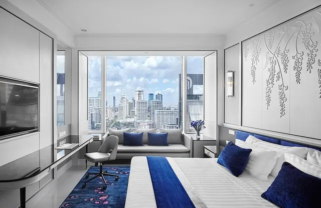 A bedroom with a made bed, a sofa by a large window looking over a city skyline, a desk with a chair, and a wall-mounted television.