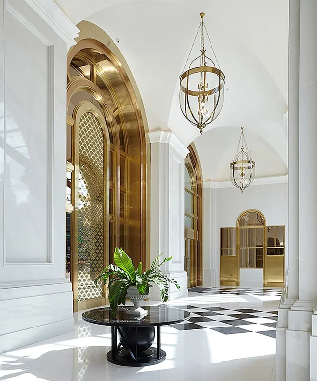 A lobby with arched gold metal screens, hanging brass lanterns, a checkered floor and a round black table holding a potted plant.