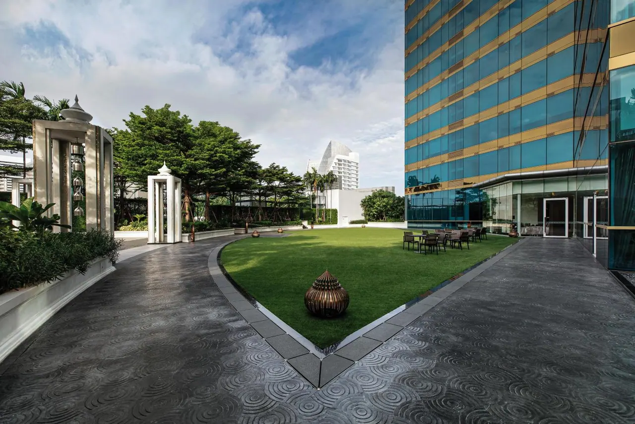 A grass lawn with a bronze urn and outdoor tables and chairs sits beside a patterned paved path and a glass-fronted hotel tower.