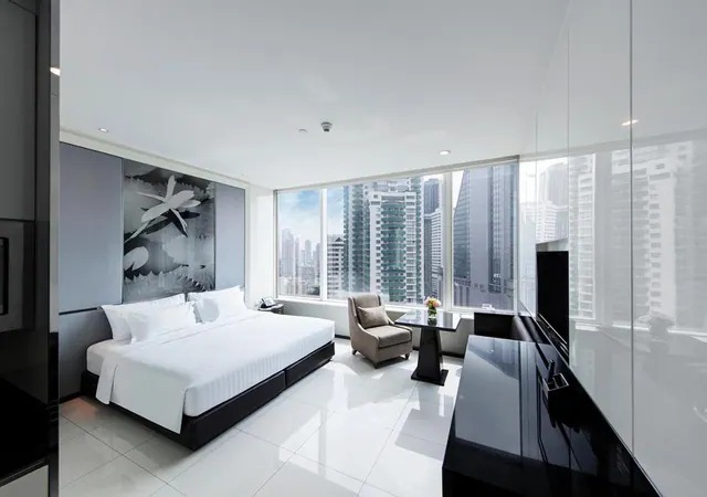 A modern hotel bedroom with a king bed, an armchair beside a small round table, a wall-mounted television, and floor-to-ceiling windows overlooking high-rise towers.