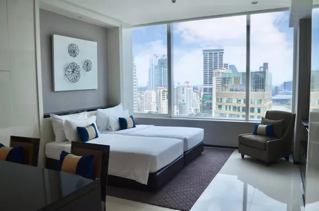 A hotel bedroom with two beds with white bedding and blue cushions, floor-to-ceiling windows showing city high-rise buildings, and an armchair beside the window.