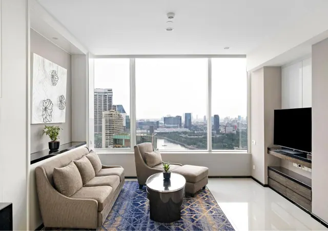 A living room with a grey sofa, armchair, ottoman and wall-mounted television, with large windows overlooking a city skyline and a river.