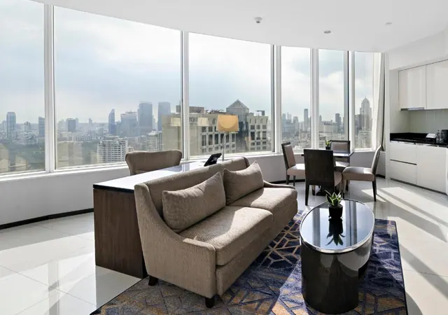 A living area with a beige sofa, a glass coffee table, a small dining table with chairs, and floor-to-ceiling windows overlooking a city skyline.
