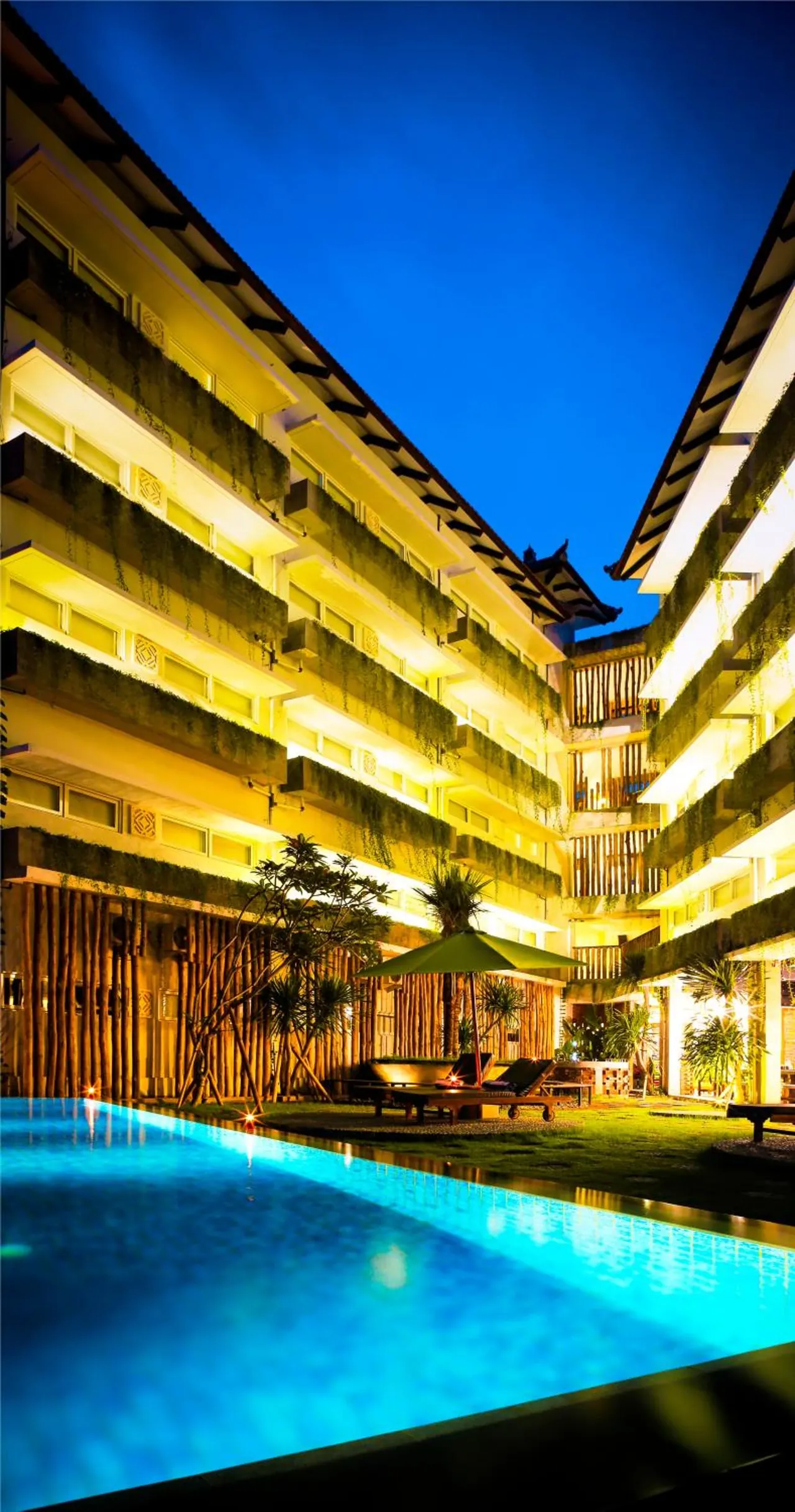 A lit swimming pool in a courtyard at dusk, with a multi-storey building with planted balconies, sun loungers and a green umbrella behind it.
