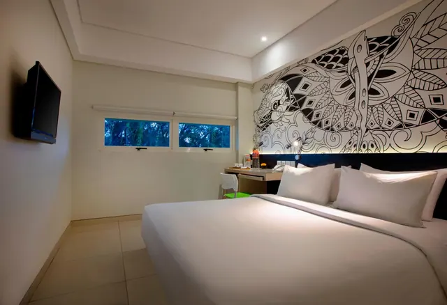 A bedroom with a made-up king bed, a black-and-white patterned wall mural, a wall-mounted television, and a desk and chair beside windows showing trees outside.