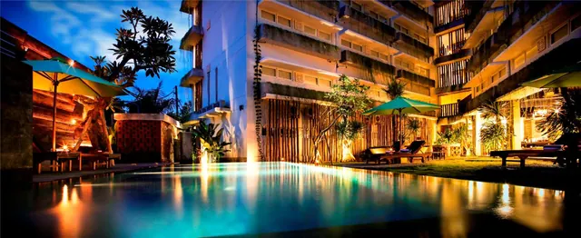 An illuminated swimming pool with sun loungers and shade umbrellas beside a multi-storey hotel building at dusk.