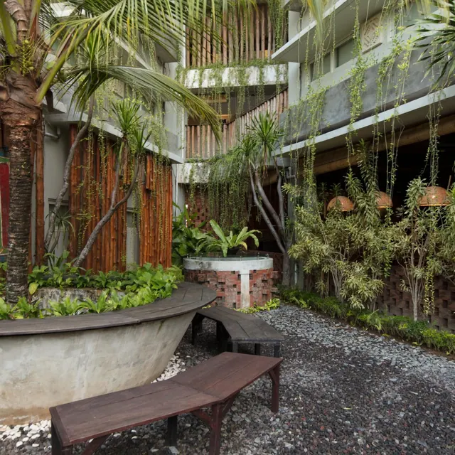 A courtyard garden with a gravel floor, wooden benches, palm and bamboo planting, and a concrete planter beside a multi-storey building.