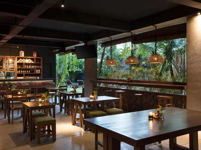 A dimly lit restaurant with wooden tables, green-cushioned stools, woven pendant lamps and a bar, with a window onto a palm garden.