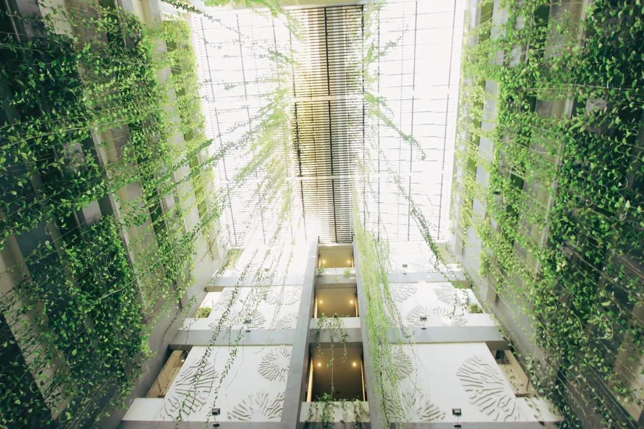 Looking up through a green-walled atrium covered in climbing vines toward a louvered skylight, with balconies visible on the lower floors.
