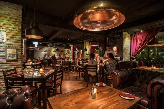 A dimly lit restaurant dining room with brick walls, copper pendant lamps, leather sofas and wooden tables, with several guests seated at tables.