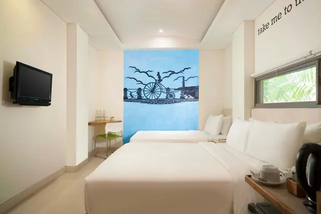 A bedroom with a large double bed and a single bed in front of a blue wall mural, a wall-mounted television, a small desk with a chair, and a window showing palm foliage.