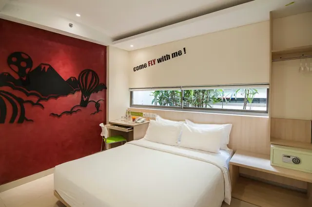 A bedroom with a double bed, a red feature wall with black cut-out mountain and hot air balloon graphics, a small desk with a chair, and an open wardrobe with a safe, with a window showing plants outside.