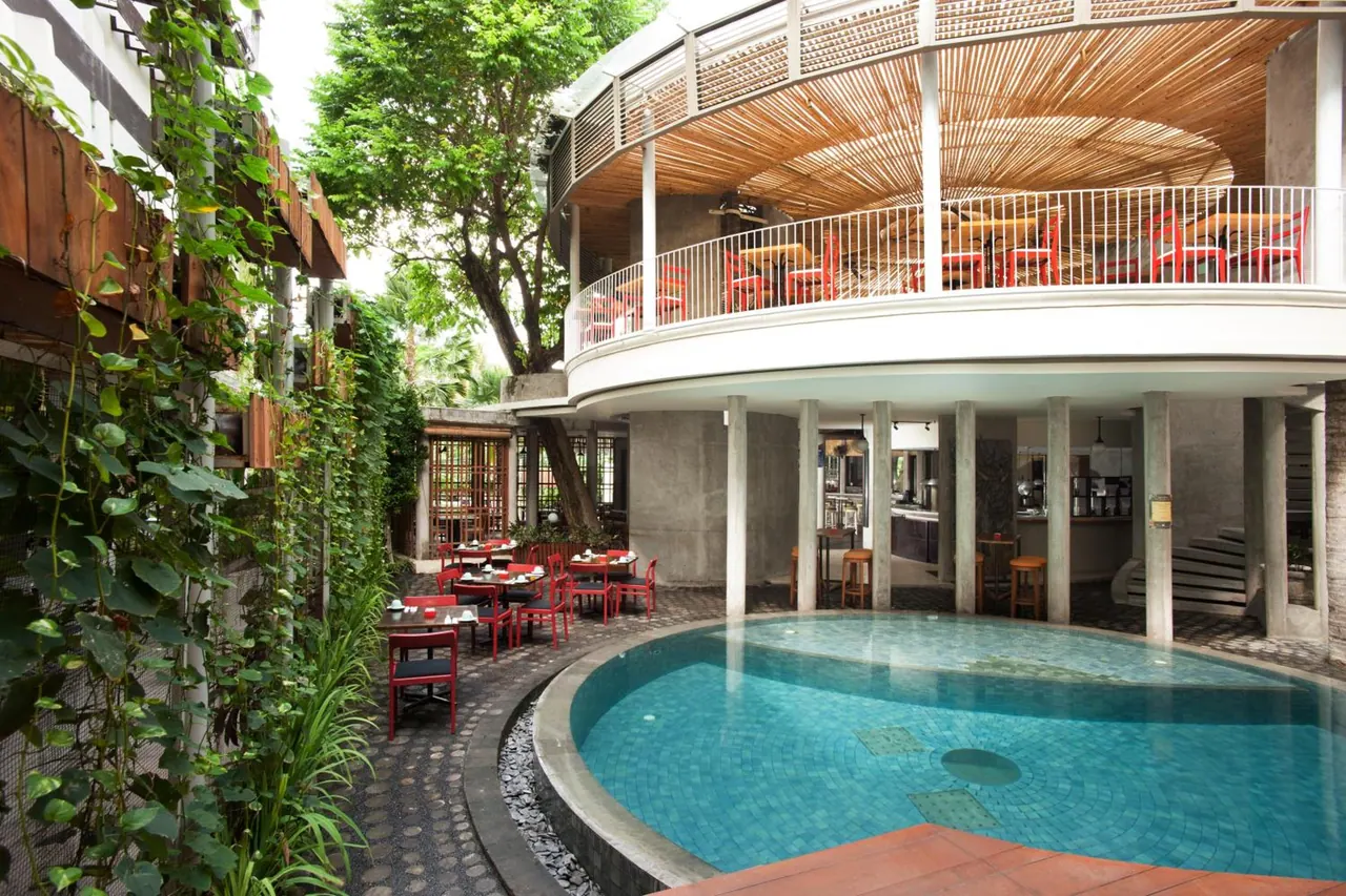 A round swimming pool sits beside an outdoor restaurant with red chairs and tables, with a bamboo-ceilinged dining terrace on the upper level.