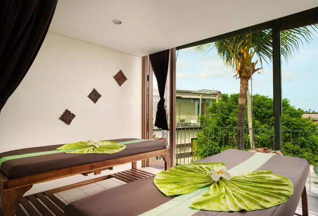 A treatment room with two massage tables dressed with folded green towels and frangipani flowers, with floor-to-ceiling windows showing a palm tree and trees outside.