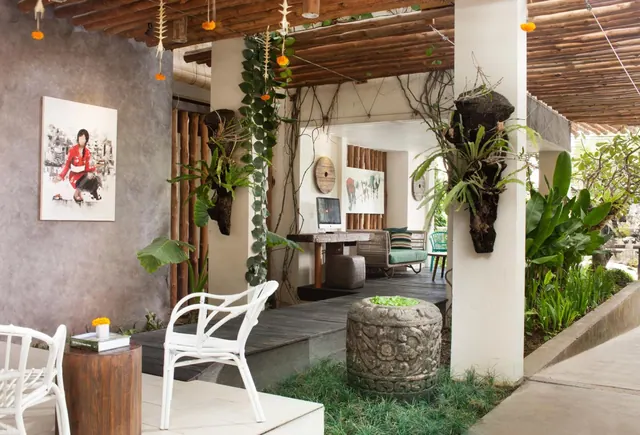 An open-air covered lounge with white chairs, a wooden side table with a book, a desk with a computer, a green-cushioned sofa, a carved stone planter and plants mounted on the pillars under a bamboo ceiling.