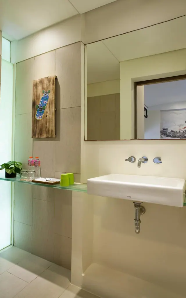 A bathroom with a wall-mounted white basin, a large mirror, a glass shelf holding a plant, water bottles and green cups, and a wooden skateboard artwork on the tiled wall.