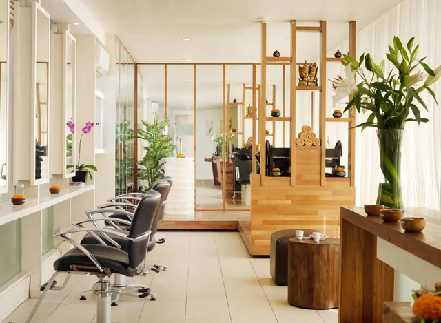 A wellness area with black styling chairs before a row of mirrors, a tall vase of lilies, and carved figures and rolled towels on a wooden partition.